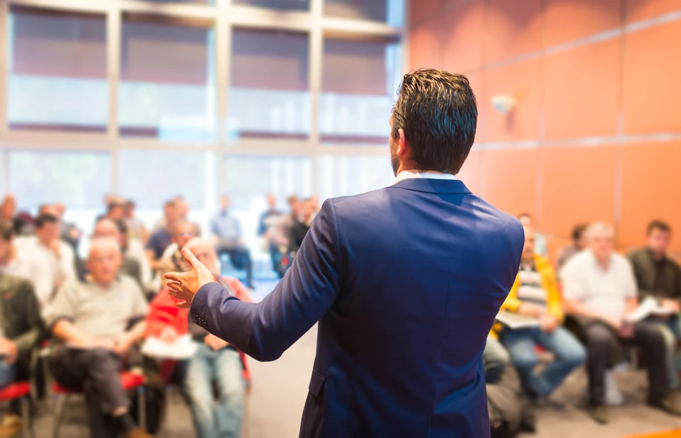 Un homme en costume bleu présente debout face à un auditoire assis dans une salle de conférence moderne
