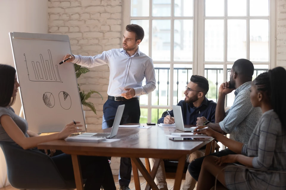 Le formateur professionnel masculin confiant présente sur des feuilles de papier à l'intention de divers participants et du personnel de l'entreprise, le chef d'équipe explique la stratégie graphique lors d'un séminaire de réunion de l'entreprise dans une salle de conseil moderne