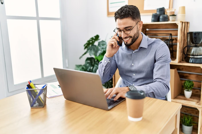 Jeune homme arabe parlant sur un smartphone utilisant un ordinateur portable travaillant au bureau