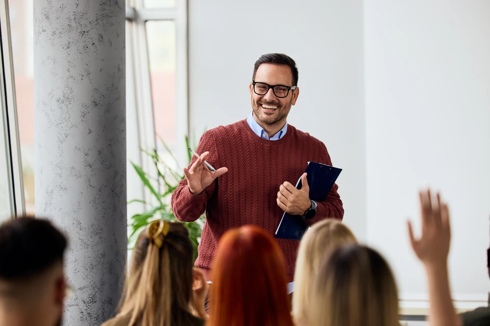 Photographie réaliste d'un groupe de participants (4-6 adultes professionnels) engagés dans une formation interactive. Ils sont assis autour d'une table ou à des postes de travail, certains consultent des écrans ou des documents, d'autres interagissent ensemble ou prennent des notes. L'atmosphère est collaborative et concentrée, avec un formateur ou un facilitateur visible en arrière-plan. L'environnement est une salle de formation moderne et accueillante, bien éclairée, avec des équipements numériques visibles (ordinateurs, tableaux). La scène illustre la mise en pratique immédiate et l'approche active mentionnées dans le contenu.